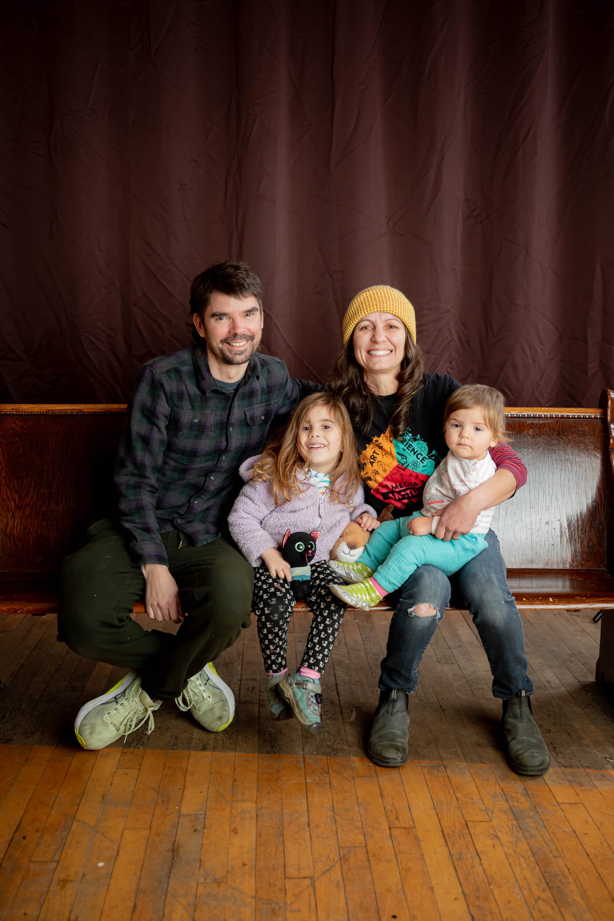 A family portrait of me, my wife Ellie, and our kids, sitting on a wooden church pew. Everyone is smiling except the youngest, she's kind of staring at the camera.