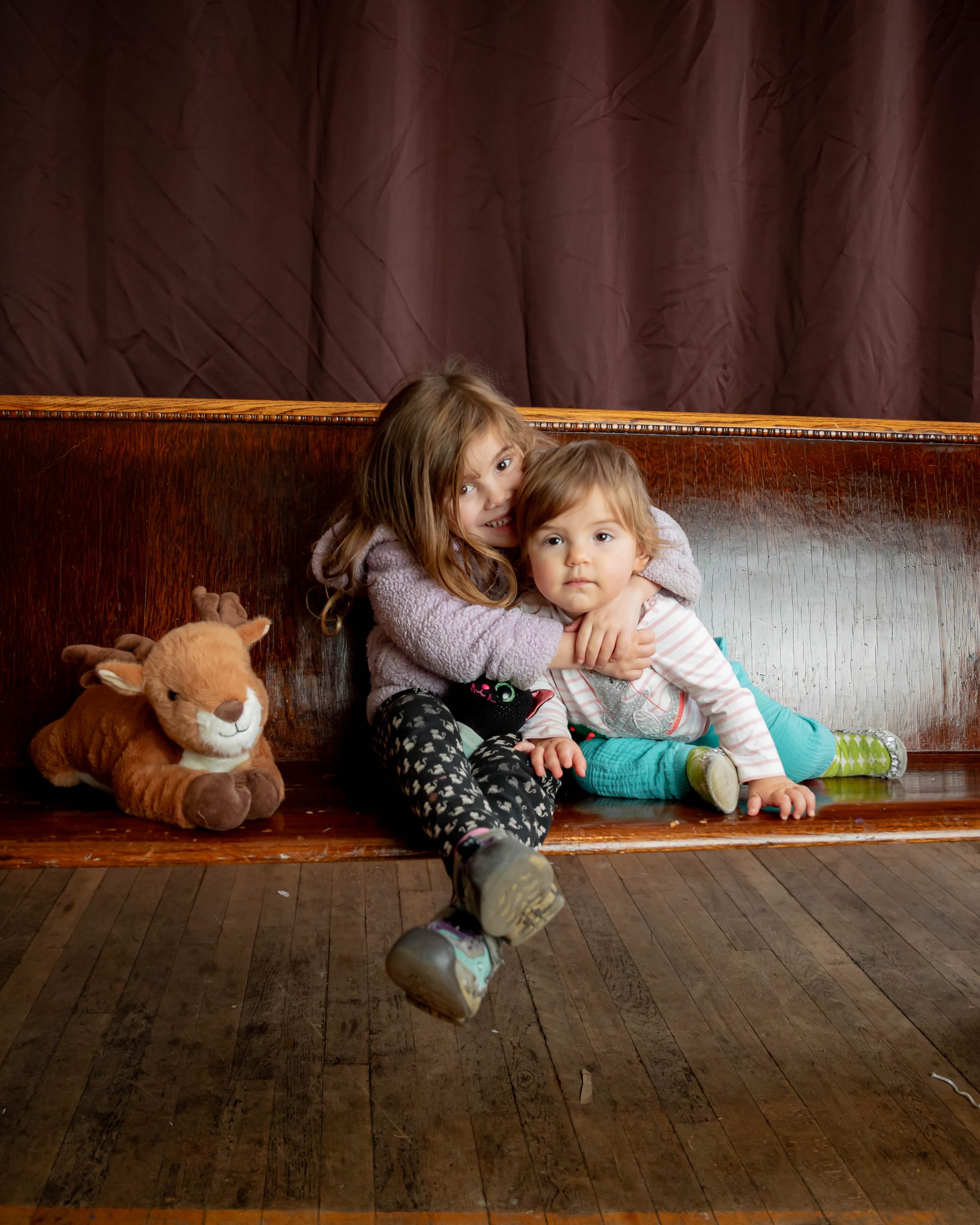 A portrait of my daughters sitting on a wooden church pew. The older one is smiling sweetly, holding the younger one in a hug that almost looks like a headlock. The younger one stares ahead, unaware of the conventions of portrait photography. A stuffed reindeer and stuffed black cat are with them on the church pew.