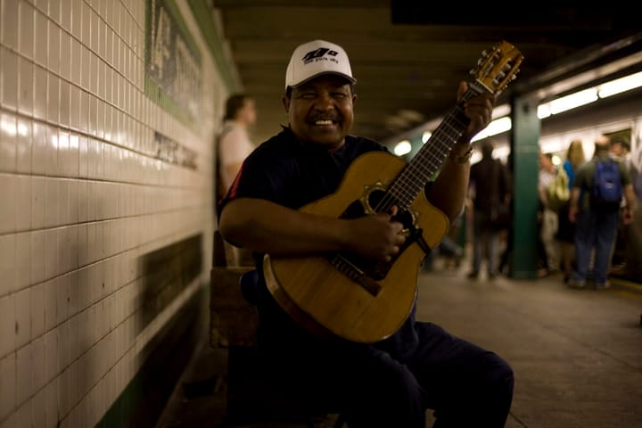 A smiling man holds a guitar on a subway platform bench.