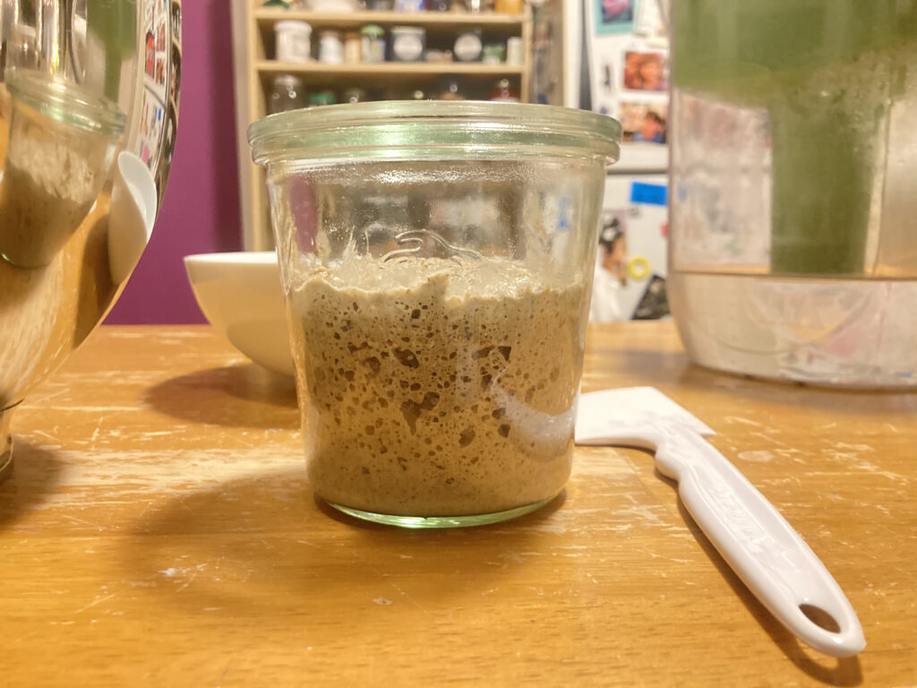 A glass jar with some sourdough starter that looks ready to bake with. It's on a countertop with various kitchen implements.
